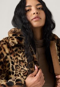 Casaco de pelúcia sintética com estampa de leopardo e fecho de correr frontal, apresentando gola e detalhes em camurça. A modelo veste uma blusa justa bege por baixo.