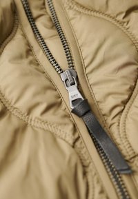 Gewatteerde jas met een tan kleurige buitenkant, voorzien van een zwarte ritssluiting en een gladde textuur, benadrukt door gestikte quiltpatronen.