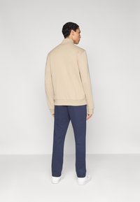 Sweat-shirt beige avec col montant, poignets et ourlet côtelés, associé à un pantalon bleu marine. Modèle portant des baskets blanches, vu de dos.