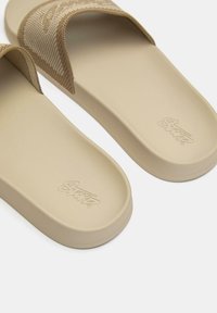 Sandali slip-on con cinturino beige testurizzato e suola beige liscia, caratterizzati da un logo impresso sulla soletta. Design minimalista.