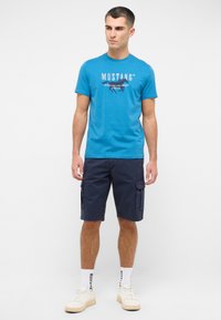 Niebieska bawełniana koszulka z logo "MUSTANG", w połączeniu z granatowymi szortami cargo. Białe trampki i wzorzyste skarpetki uzupełniają strój.