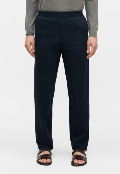 Homme portant un pantalon droit bleu marine foncé, un haut gris à manches longues et des sandales en cuir marron, debout devant un fond neutre.