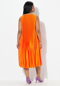 Vestido naranja plisado con cuello alto y diseño sin mangas, que presenta una silueta fluida. Combinado con zapatos de tacón alto morados.