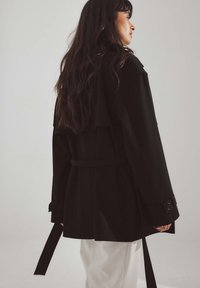 Cappotto nero lungo con schiena a soffietto, vita con cintura e polsini abbottonati, caratterizzato da una vestibilità sagomata e una texture morbida. Pantaloni bianchi visibili sotto.