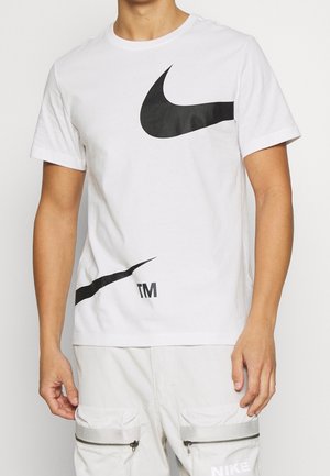 Mężczyzna w białym T-shircie Nike z dużymi czarnymi logo Swoosh i białych spodniach cargo z zamkami błyskawicznymi.