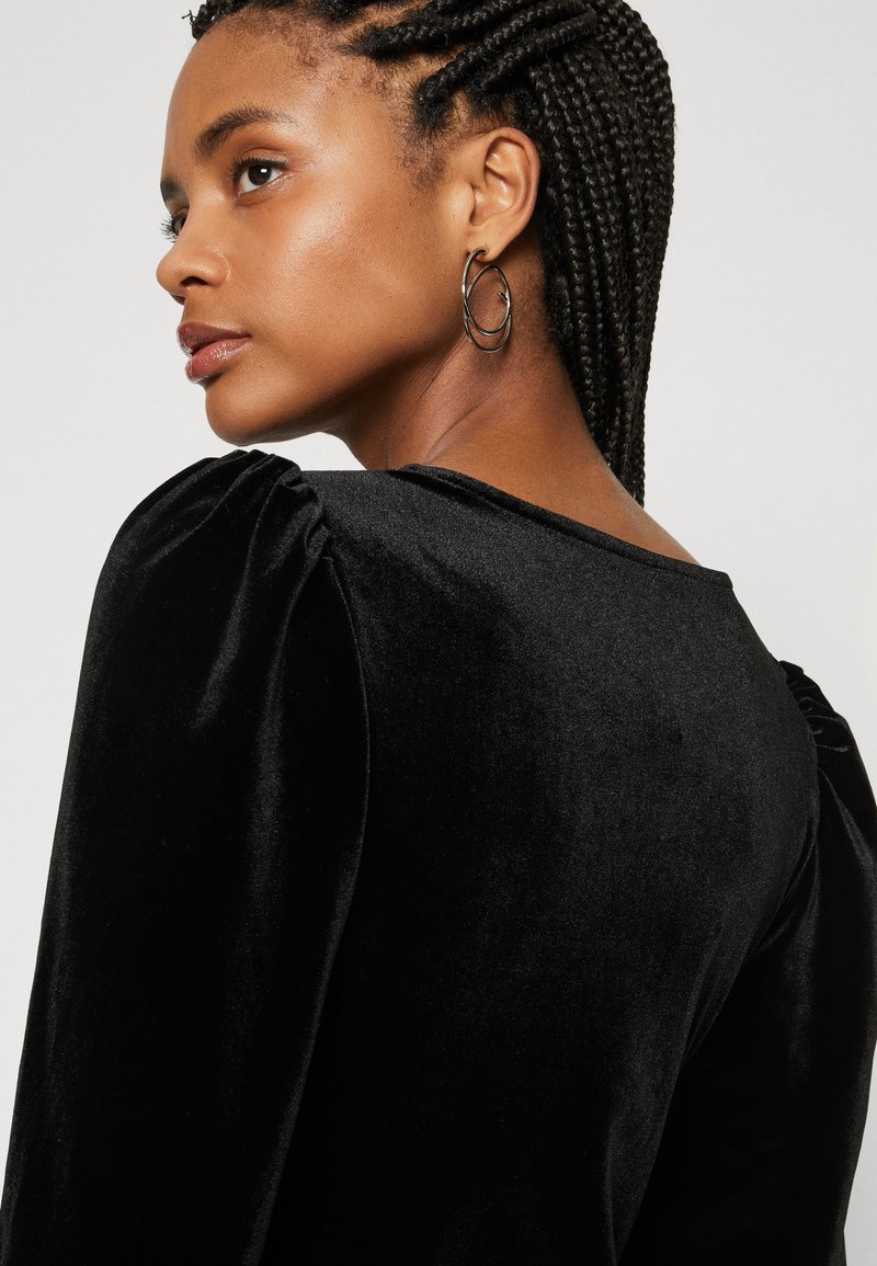 Schwarzes Samtoberteil mit puffierten Ärmeln und weitem Ausschnitt. Akzentuiert mit minimalistischen Creolen und geflochtenem Haardetail.