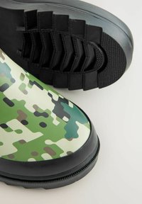 Bottes imperméables vertes et beige avec un motif camouflage, dotées d'une semelle extérieure en caoutchouc noir texturé et d'un design incurvé.