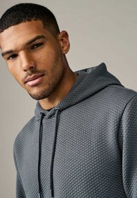 Grijze hoodie van een gestructureerde, diamantpatroon stof. Voorzien van een trekkoordhoed en een relaxte fit, met een close-up van de halslijn.