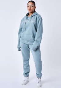 Un sweat à capuche bleu clair avec des cordons de serrage et une poche avant, associé à un pantalon de survêtement assorti. Le tissu semble doux avec des accents côtelés. Un texte de logo blanc est visible.