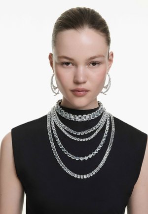 Des colliers en crystal transparent multi-couches de différentes formes et tailles, associés à des boucles d'oreilles créoles en crystal. Porté sur un haut noir.