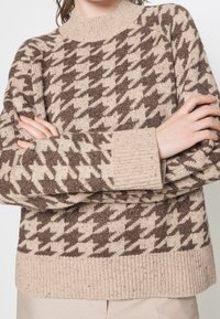 Beige und braun karierter Strickpullover im Hahnentrittmuster mit gerippten Bündchen und Saum, getragen von einer Person mit verschränkten Armen.