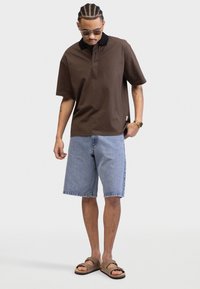 Brown cotton polo shirt with a black collar, paired with light blue denim shorts. Worn with brown sandals and sunglasses.