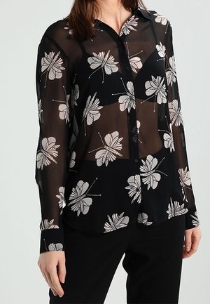 Chemise noire transparente avec un motif floral blanc, manches longues, devant à boutons et coupe ample. Détails notables incluant un col évasé et une texture contrastante.