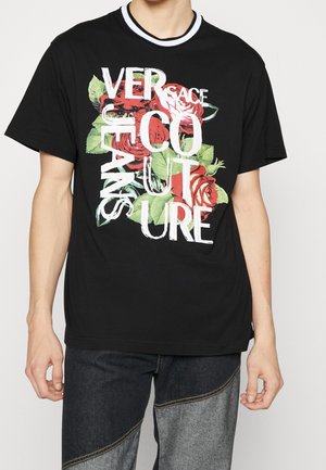 Personne portant un t-shirt noir avec un motif floral blanc et rouge et un texte blanc superposé indiquant "Versace Jeans Couture".