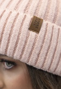 Berretto a maglia rosa con texture a coste, dotato di un'etichetta in pelle marrone con la scritta "VIVID SENCE" in rilievo. Focus sul primo piano del cappello.