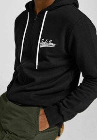 Sort zip-up hoodie lavet af blødt stof, med hvide snore og logo broderi. Designet inkluderer en kængurulomme og ribbede manchetter.