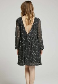 Robe noire fluide avec de longues manches transparentes, présentant un dos en V profond et un motif floral blanc éparpillé sur l'ensemble. Détails de tissu texturé visibles.