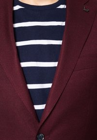 Burgundowa blazer z fakturowanej tkaniny, z klapami, czarnym guzikiem, nałożona na niebiesko-białą koszulkę w paski, z akcentem na kontrastujące kolory i materiały.