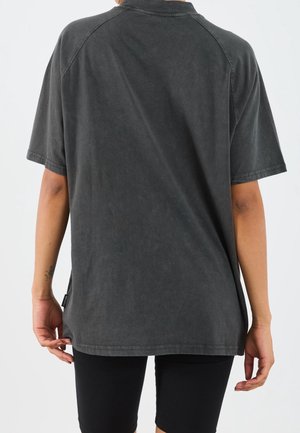 Personne portant une chemise ample à manches courtes gris foncé et un short noir, vue de dos sur un fond uni.