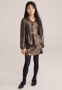 Metāla bronzas kleita ar pepluma apakšmalu, garām piedurknēm un v-veida dekoltē. Kombinēta ar melnajām zeķbiksēm un spīdīgām melnām kurpēm.