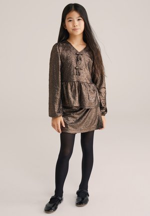 Metāla bronzas kleita ar pepluma apakšmalu, garām piedurknēm un v-veida dekoltē. Kombinēta ar melnajām zeķbiksēm un spīdīgām melnām kurpēm.