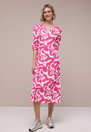 Vrouw staat en lacht, draagt een roze-witte midi-jurk met een abstract patroon, opgerolde mouwen en zilveren sneakers, tegen een neutrale achtergrond.