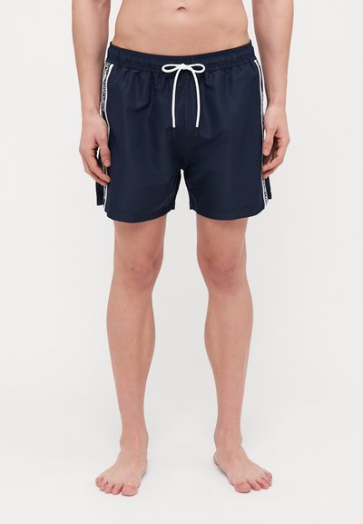 Pantaloni da nuoto blu navy con cintura elastica e coulisse bianca. Le strisce laterali presentano un motivo a contrasto. Tessuto liscio e leggero.