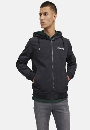 Black hooded jacket with zip closure, featuring side pockets and ribbed cuffs. Brand logo visible on the chest. Layered with a green hoodie.