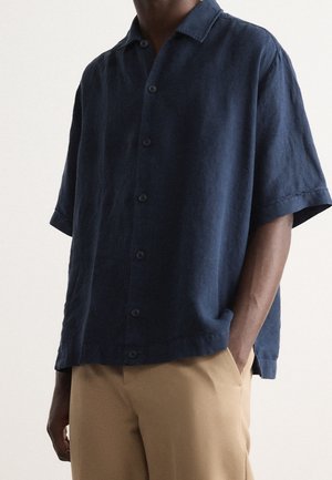 Mężczyzna ubrany w luźną, krótko-rękawową granatową koszulę na guziki oraz beżowe spodnie, stojący z jedną ręką w kieszeni.