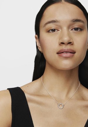 Collar de pendant de plata en una modelo, que presenta un diseño circular y una cadena fina, llevado con una blusa negra sin mangas. Textura suave.