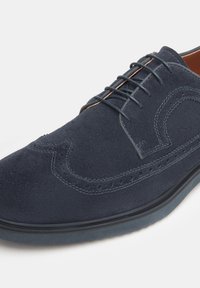 Chaussure à lacets en suede navy avec des détails perforés, bout rond et accents en cuir marron contrastant sur les côtés et la languette.