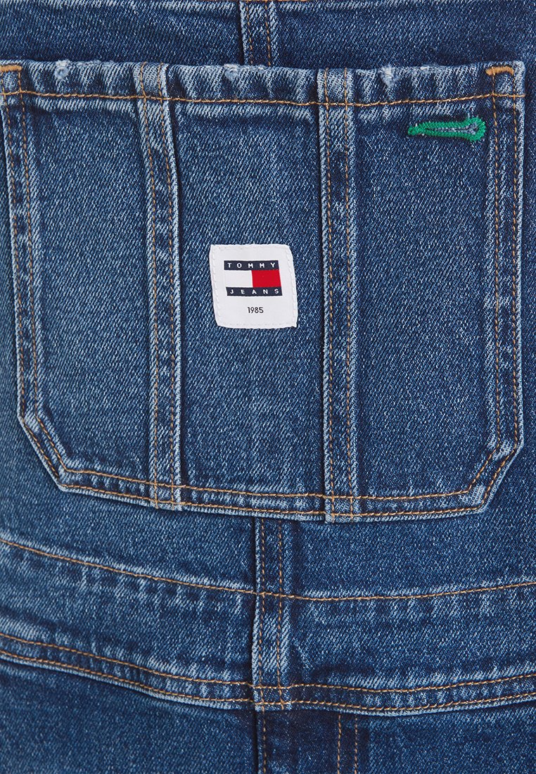 Salopette en denim avec une poche arrière portant une étiquette blanche affichant un texte de logo rouge, blanc et bleu. Détails usés et couture jaune.