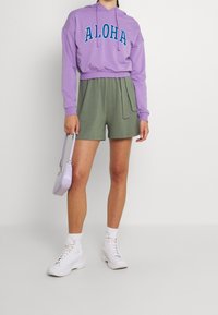 Haut de survêtement court violet avec le texte "ALOHA", shorts verts avec cordon de serrage, bottes blanches et un petit sac à main lavande.