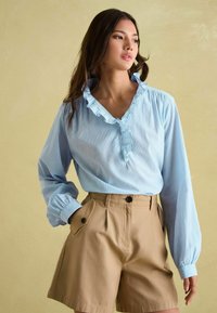 Svetlomodrá pruhovaná blúzka s volánikmi okolo goliera a dlhými rukávmi, skombinovaná s khaki vysokými šortkami s gombíkovými vreckami.