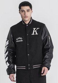 Čierna varsity bunda s vlneným telom a umelými koženými rukávmi. Obsahuje výšivku "K" a nášivku s nápisom "GIANNI KAVANAGH". Prúžkované rebrové manžety.