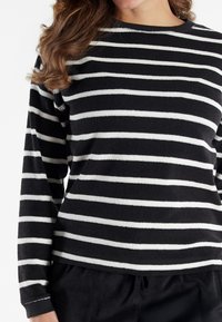 Frau in einem schwarzen Langarmpullover mit gleichmäßig verteilten horizontalen weißen Streifen und schwarzen Hosen.