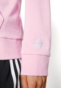 Różowa bluza z ribbowymi mankietami, z małym białym haftowanym logo Adidas na rękawie. Materiał wydaje się miękki i teksturowany.