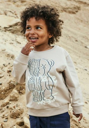 Smilende barn med krøllete hår iført beige genser med grafisk bjørnemotiv, stående utendørs på sandete underlag.