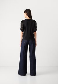 Μαύρη μπλούζα με ανθεκτικά μανίκια από δαντέλα, σε συνδυασμό με σκούρα μπλε παντελόνα. Απλός σχεδιασμός με χαλαρή εφαρμογή και ελάχιστες λεπτομέρειες.