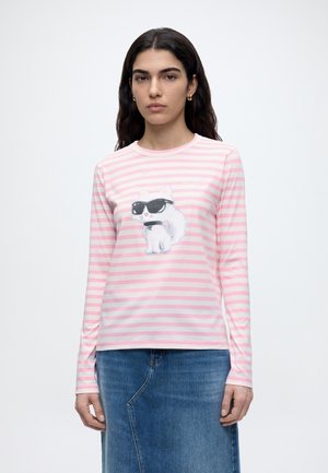 Νεαρή γυναίκα με σκούρα μαλλιά φοράει ροζ και λευκό μπλουζάκι με ρίγες μακριάς μανσέτας που έχει μια λευκή γάτα με γυαλιά ηλίου και ένα τζιν φούστα.