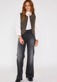 Un jean gris évasé associé à un gilet texturé de couleur marron foncé avec un motif en chevrons et des boutons noirs, porté par-dessus une blouse blanche.