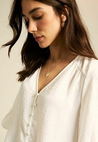 Witte gekreukte stoffen blouse met een V-hals, voorzien van knopen aan de voorkant en lange mouwen, gecombineerd met gelaagde gouden kettingen en oorringen.