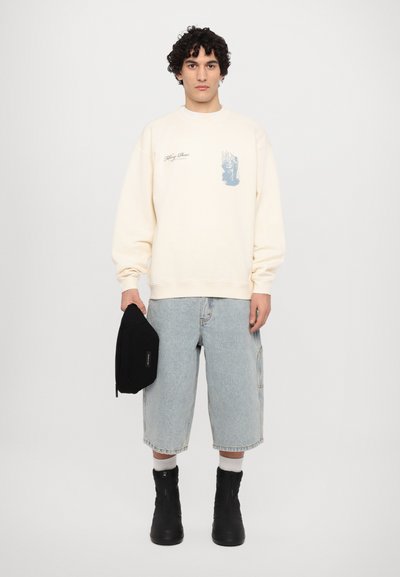 Cremefarbener Sweater mit grafischem Design, kombiniert mit hellblauen Jeansshorts. Dazu eine schwarze Tasche und schwarze Ankle Boots.
