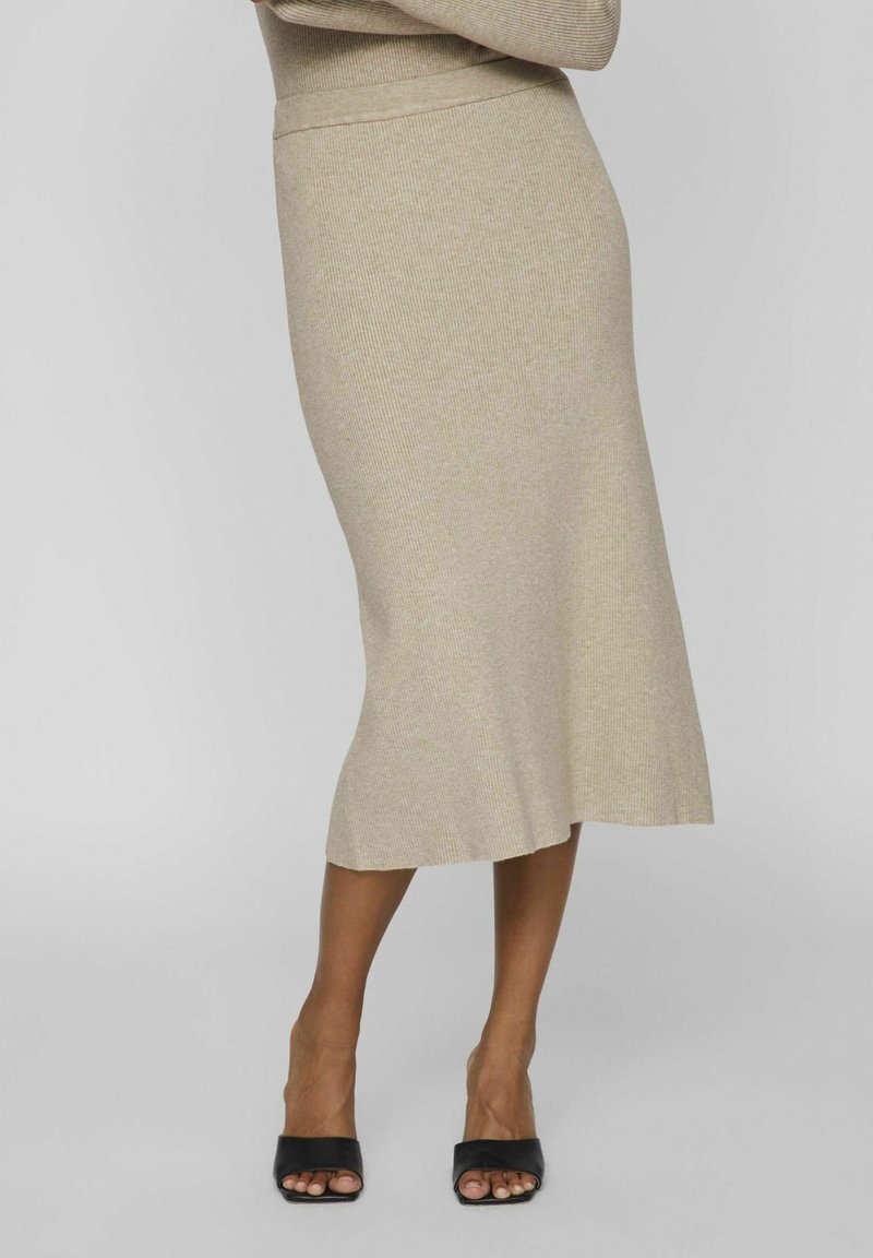 Femme portant une jupe midi côtelée beige associée à des sandales à talons ouvertes noires sur un fond gris uni.