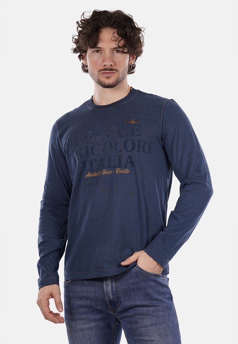 Uomo con capelli ricci che indossa una camicia a maniche lunghe blu navy con il testo "Frecce Tricolori Italia" e jeans blu, in piedi con una mano in tasca.