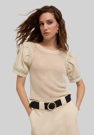 Beige nettop met opstaande korte mouwen en een ronde halslijn. Gepaard met hooggerokte crèmekleurige broek en een zwarte riem.