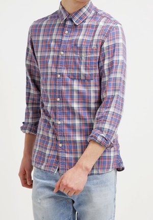 Mann trägt ein rot-weiß-blau kariertes Hemd mit hochgekrempelten Ärmeln und hellen, verwaschenen Jeans vor einfachem Hintergrund.