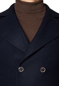 Granatowy wełniany płaszcz o podwójnym zapięciu, z klapami i metalowymi guzikami, noszony na brązowym, prążkowanym swetrze z golfem.