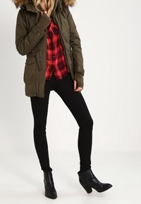 Osoba v parce se zeleným kožíškem na okrajích, červené kostičkované košili, černých slim kalhotách a černých bot�ách na podpatku, stojící s jednou nohou mírně pokrčenou.