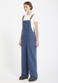 Denim salopettes in medium blauw, met wijde benen, verstelbare bruine banden en voorkwasten. Gecombineerd met een aansluitend wit t-shirt.
